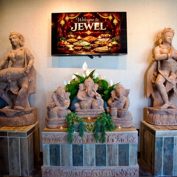 Indian-themed decor with statues of deities, plants, candles, and a TV displaying "Welcome to JEWEL" with food.
