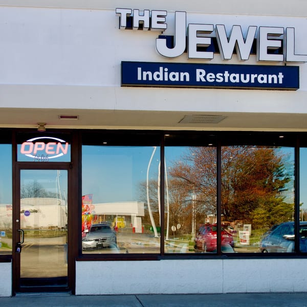 The Jewel Indian Restaurant storefront with an "OPEN" sign and reflections of cars and trees in its windows.
