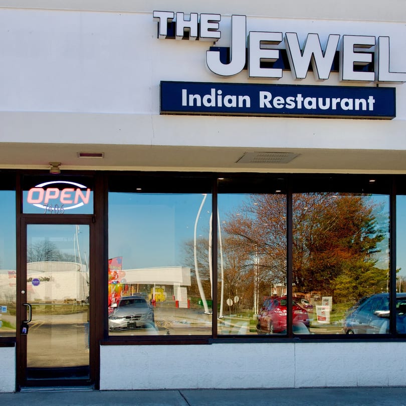 The Jewel Indian Restaurant storefront with an "OPEN" sign and reflections of cars and trees in its windows.