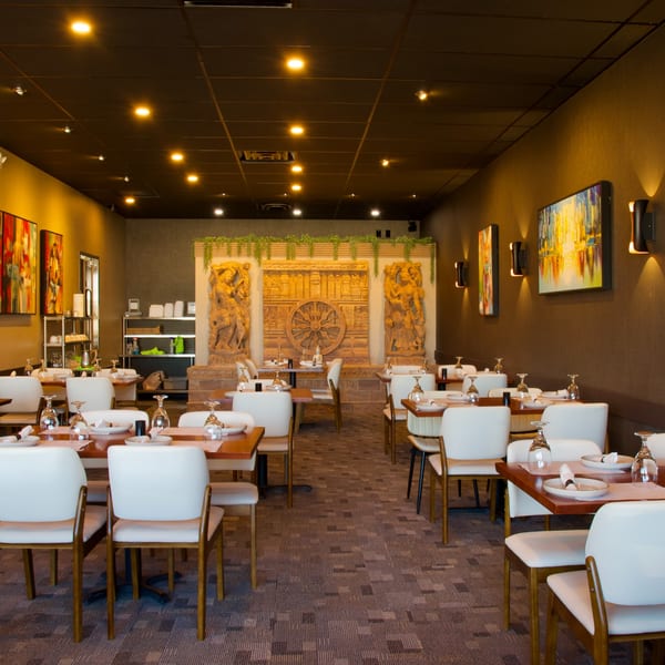 Empty restaurant dining room with white chairs, wooden tables, and ornate wall carvings.