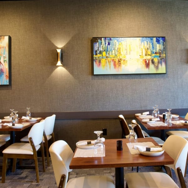 A modern restaurant dining room with set tables, white chairs, and abstract art on textured walls.