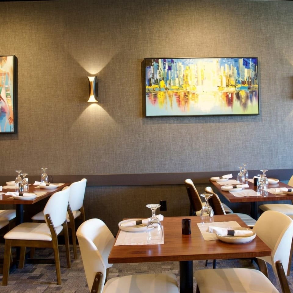 A modern restaurant dining room with set tables, white chairs, and abstract art on textured walls.