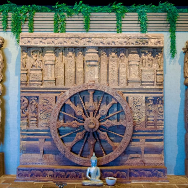 Intricately carved wooden wall with a large wheel, a small Buddha statue, and green vines.