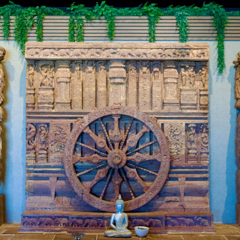 Intricately carved wooden wall with a large wheel, a small Buddha statue, and green vines.