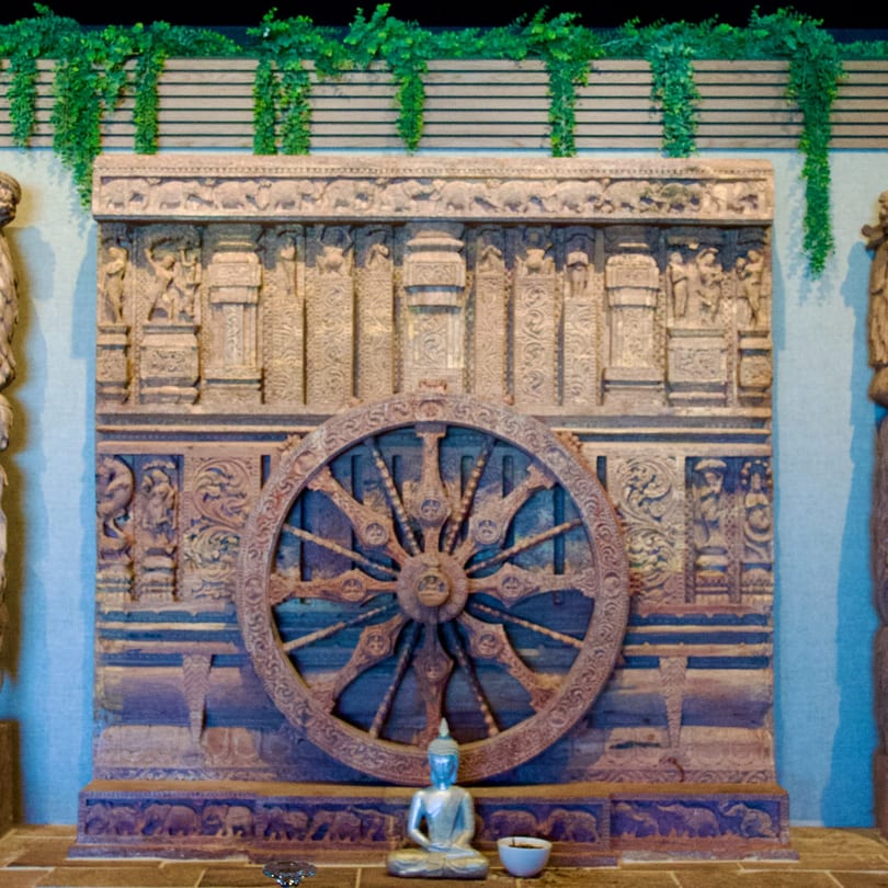 Intricately carved wooden wall with a large wheel, a small Buddha statue, and green vines.
