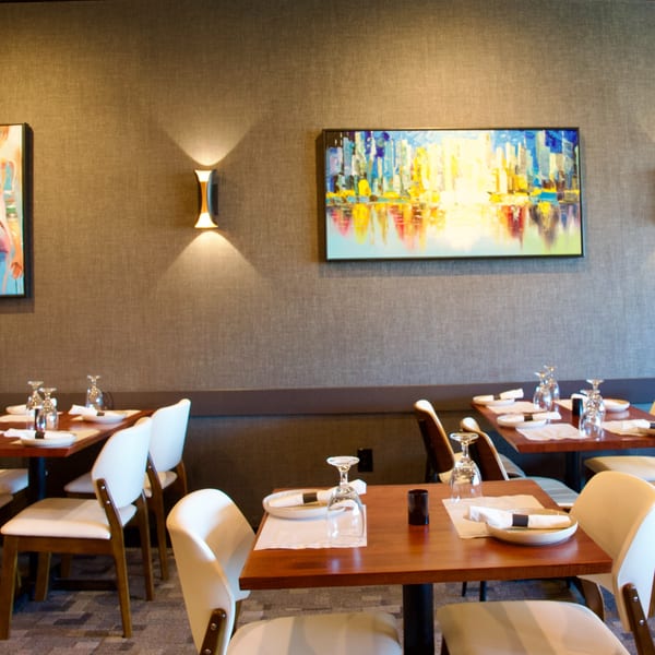 A modern restaurant dining room with set tables, white chairs, and abstract art on textured walls.