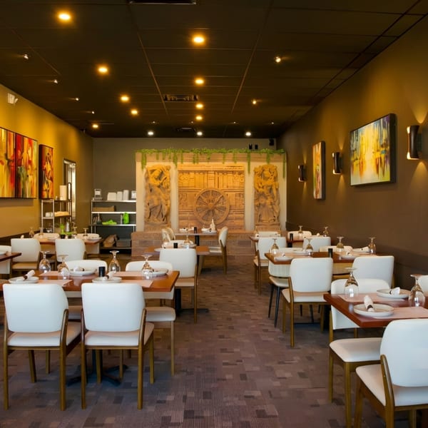 A restaurant dining room with white chairs, wooden tables, and decorative wall carvings under warm lighting.